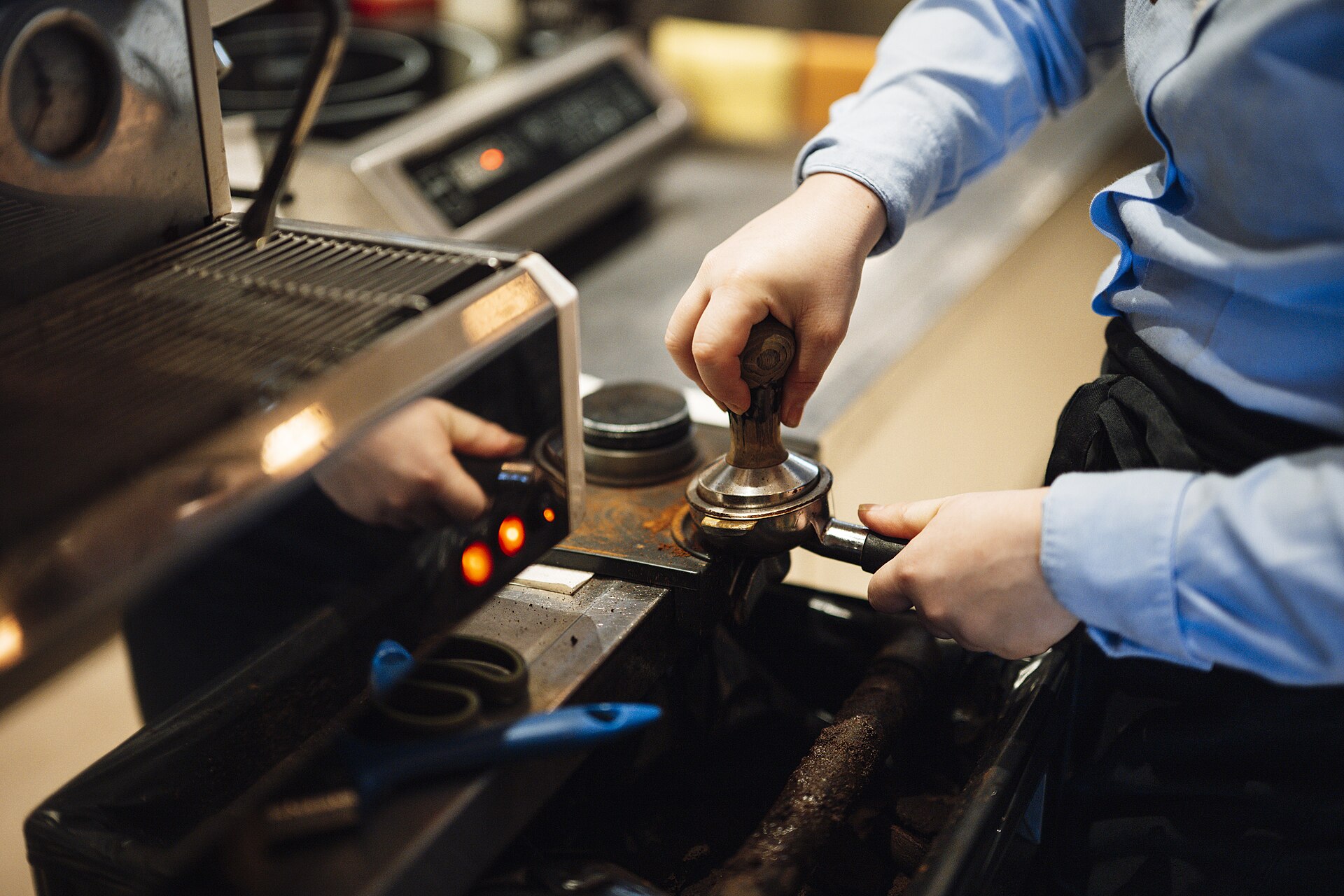 A barista tamping fresh coffee into a portafilter next to the espresso machine.