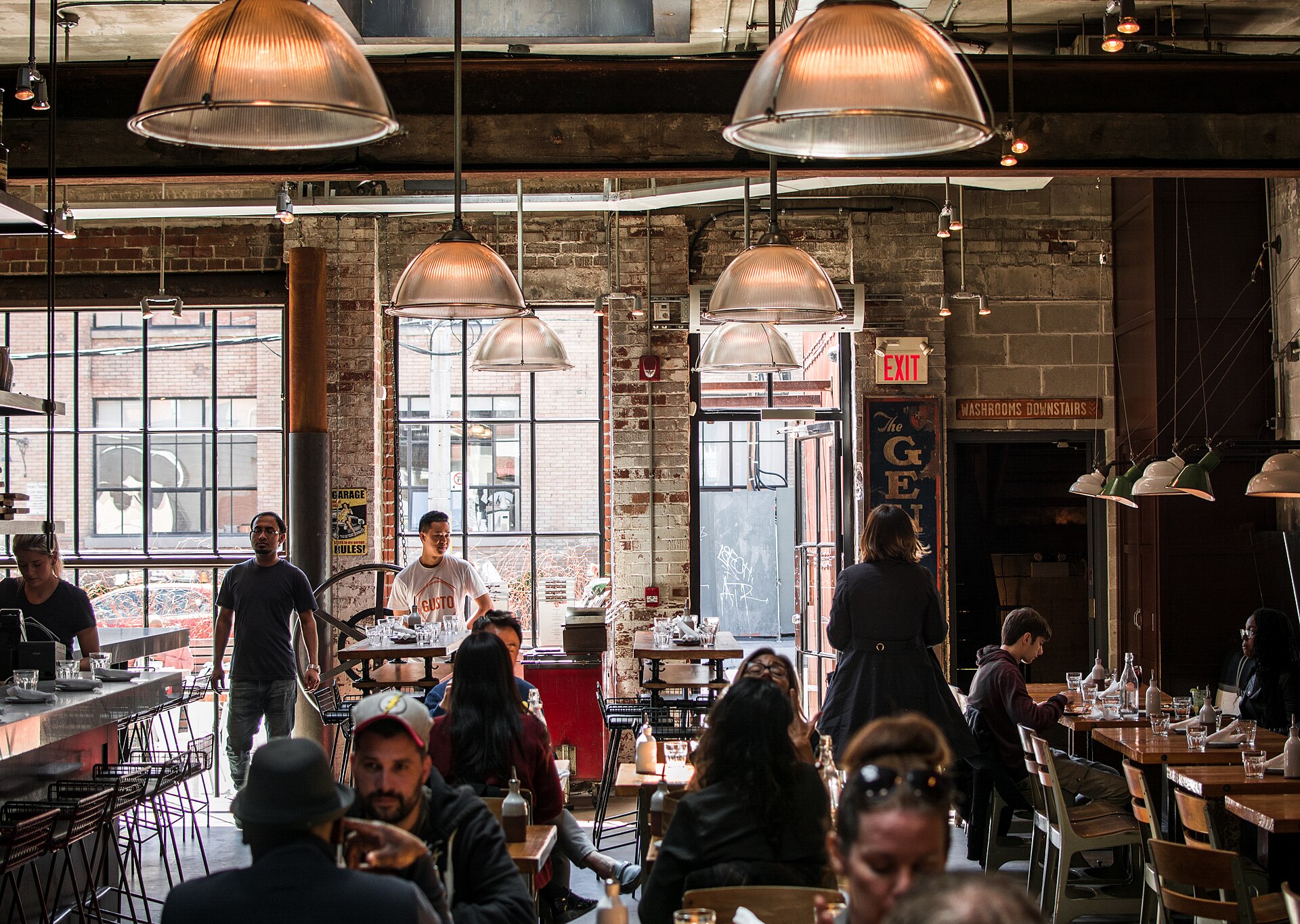 A busy café with brick walls, big factory windows and pendant lamps, guests talking at wooden tables.