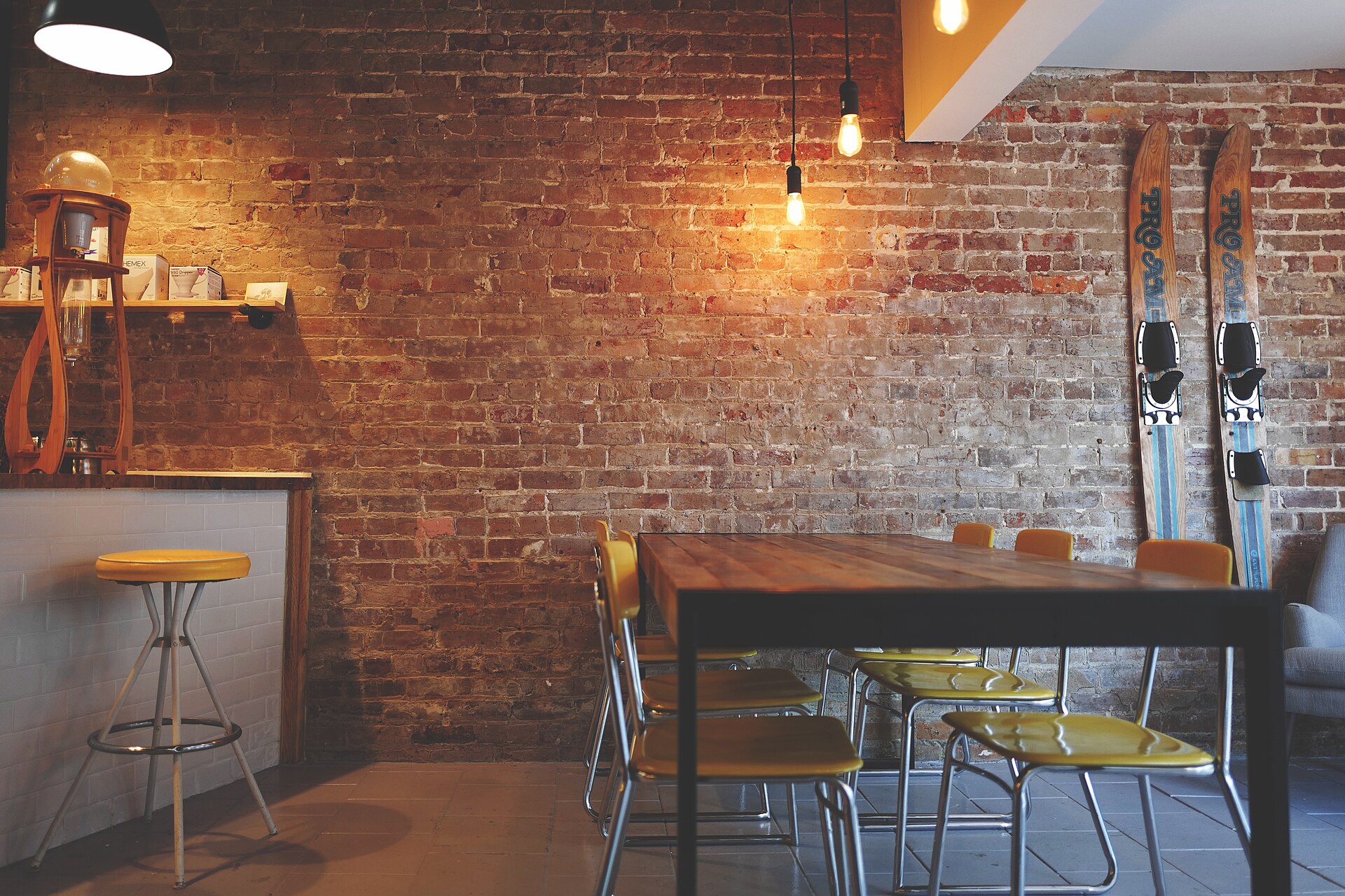 A long wooden table with yellow chairs against a brick wall, lit by bare bulbs.